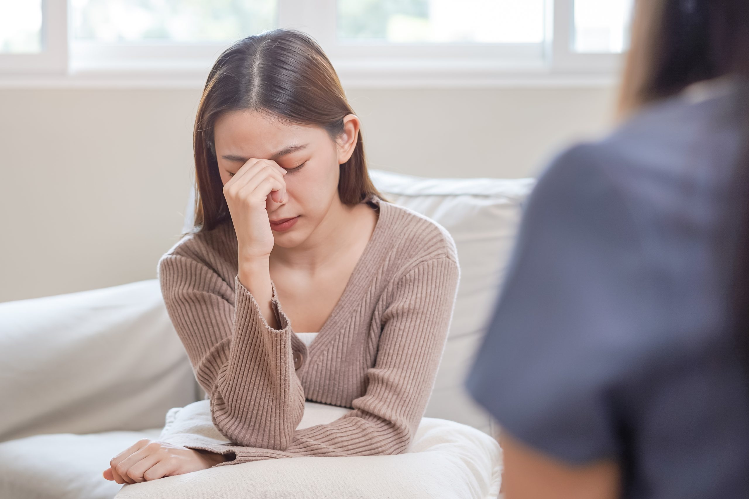 Psychology, depression. Sad, suffering asian young woman consult A young woman sits on a sofa with her head resting in her hand, eyes closed in distress. She appears to be in a therapy session, facing a counsellor who is slightly blurred in the foreground. The image captures a moment of emotional pain, possibly related to anxiety or depression, in a calm indoor setting.